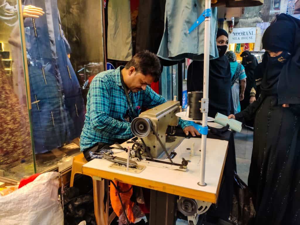 Tailor stitches Burkhas outside a store as women watch