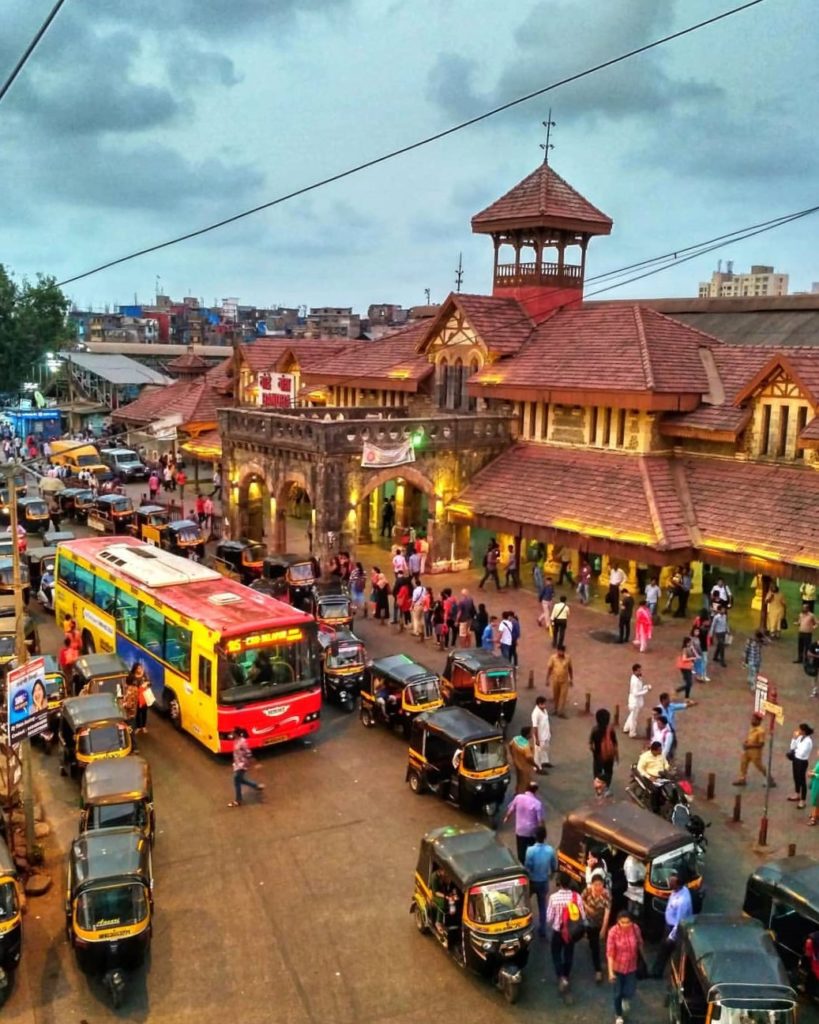 The Bandra West railway station area in the evening, with the structure lit up by lights and rickshaws, a bus and commuters scattered