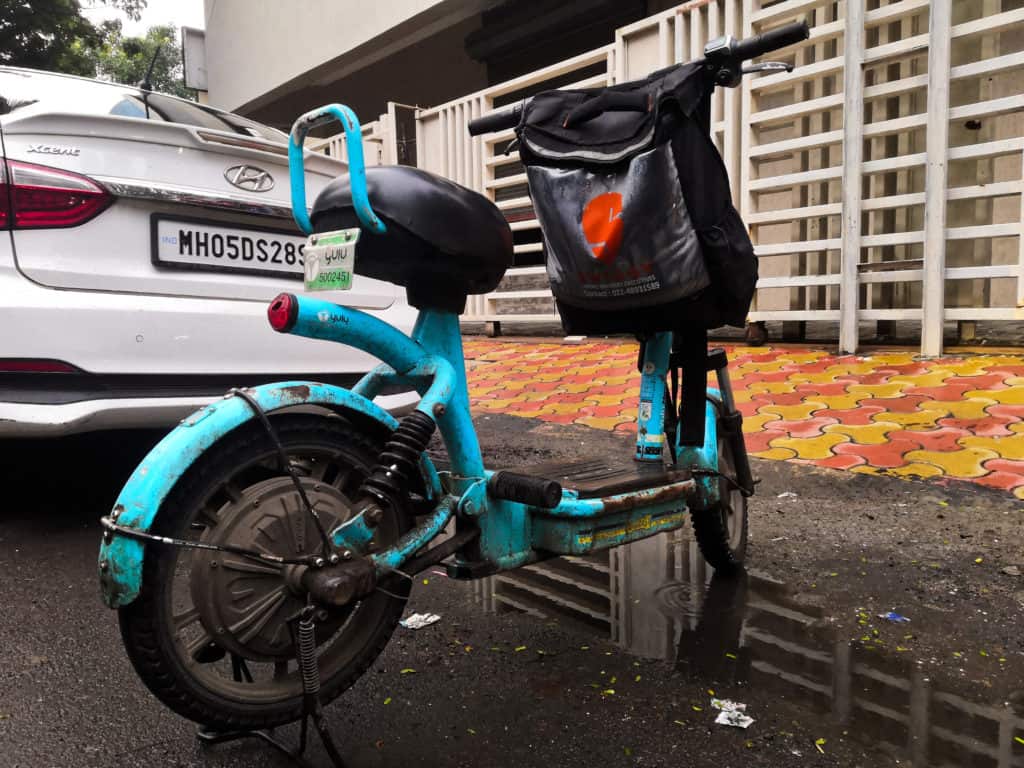 A blue electric moped bike with a Swiggy carry bag on the handlebars