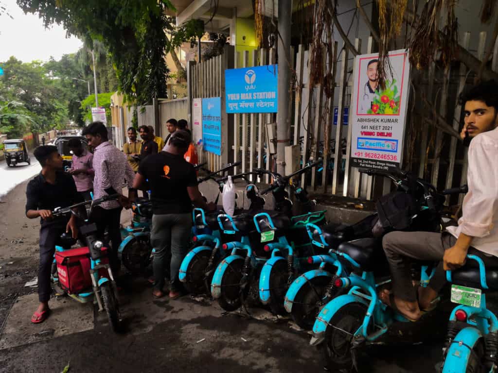 A row of delivery workers lined up in blue electric mopeds in front of a charging station 