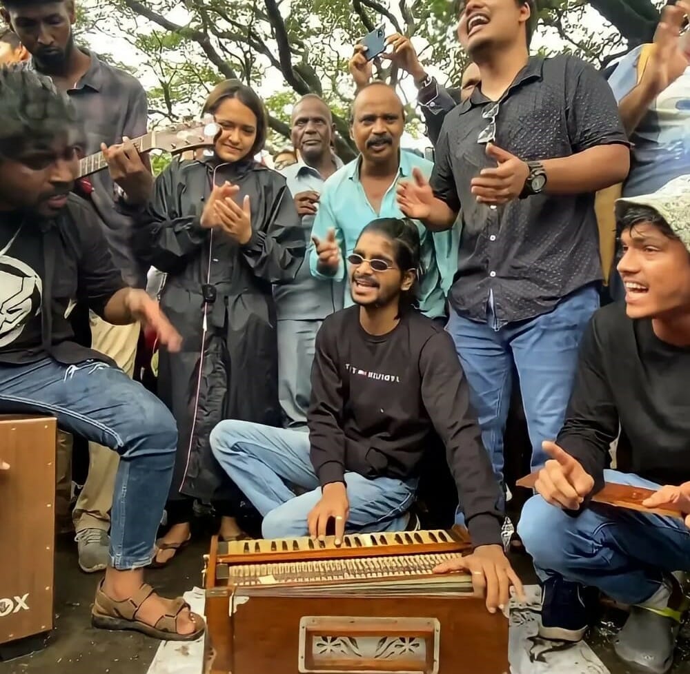 Members of Aarey Key Foundation are performing for protestors at Aarey. The man in the middle is playing harmonium and the one next to him is playing the cajon. There are a lot of people around them who are singing and clapping along.  