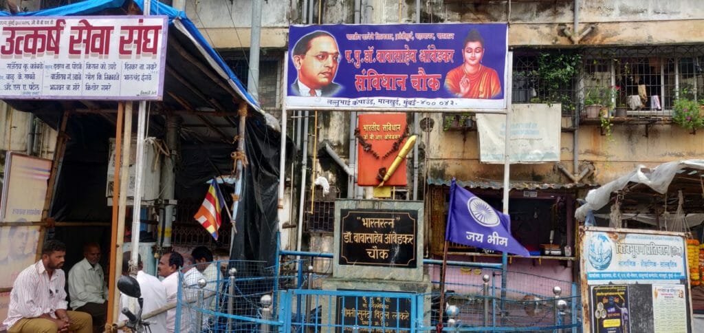 There is a banner with a photo of Dr BR Ambedkar and a plank that reads, "Bharat Ratna Dr Babasaheb Ambedkar Chowk". Beside that is a dark blue flag with Ashok Chakra in the middle and the words Jai Bhim written below it.  