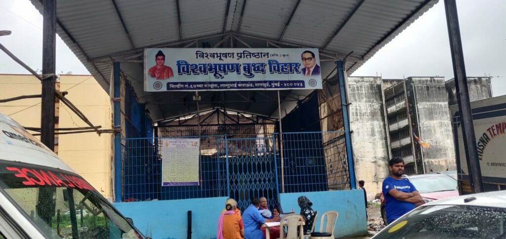 People are seen sitting on the chairs put outside Buddha Bihar. There is a banner with Gautam Buddha's picture on the left and Dr Br Ambedkar's picture on the right.