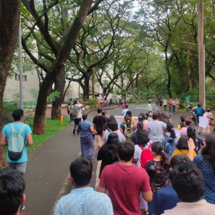 A large group of students is seen marching in protest with a banner within IIT Bombay campus. The picture is captured by one of the protestors from the back and none of their faces are visible.  