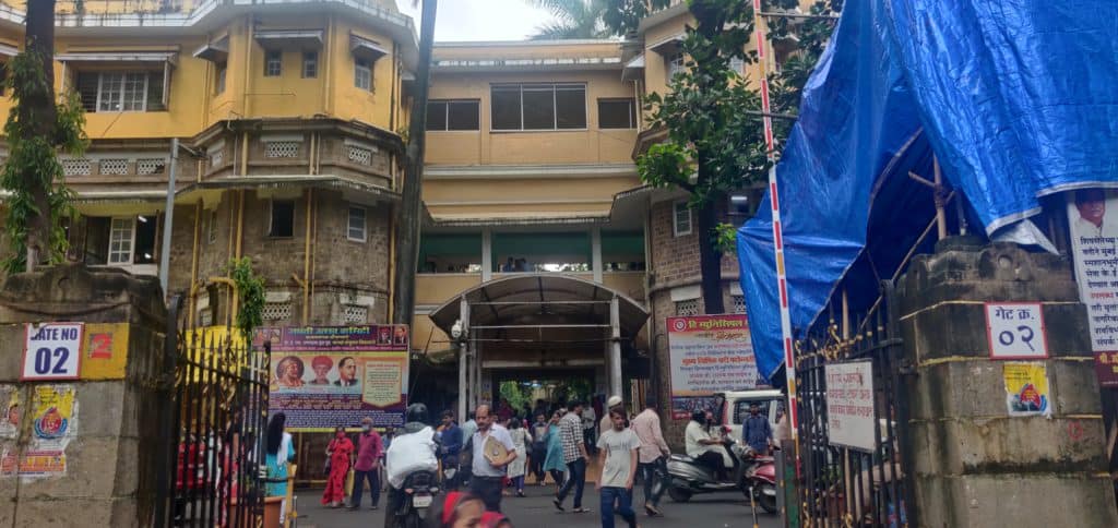 An image of KEM hospital's main gate. A crowd is walking around within the hospital premises, navigating through the space.