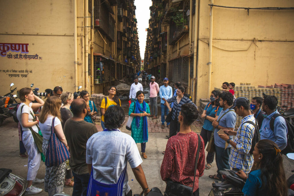 About 10 to 15 people are see standing in a circle in front of a SRA building at Lallubhai Compound. The participants are mostly young and are being briefed about the walk. 