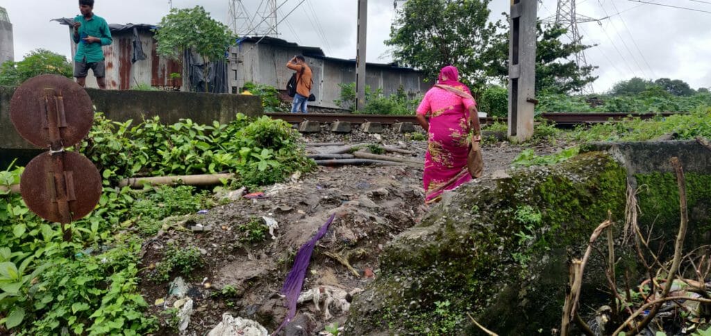 A middle aged woman, wearing a saree, her back facing the camera is seen walking towards the railway tracks on a dilapidated road. 