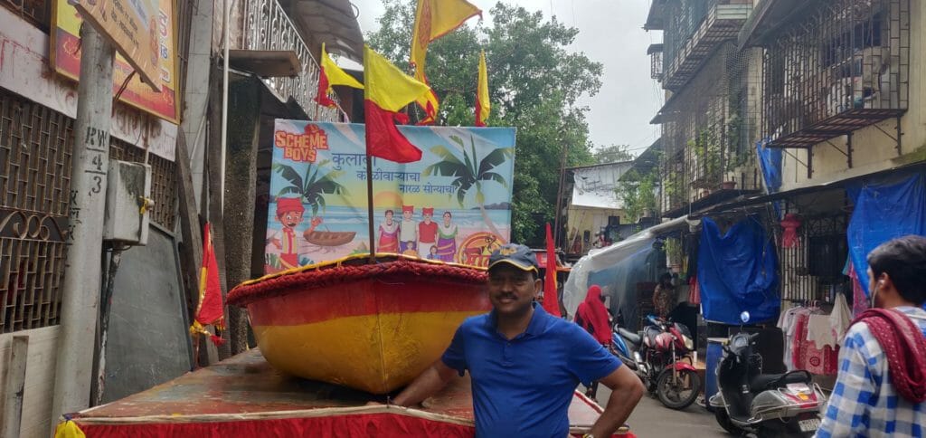 Paresh Sirtandel standing in front a boat at Coloba Koliwada. The boat will be used in Narali Purnima celebrations. Sirtandel is wearing a blue t-shirt and a cap. 