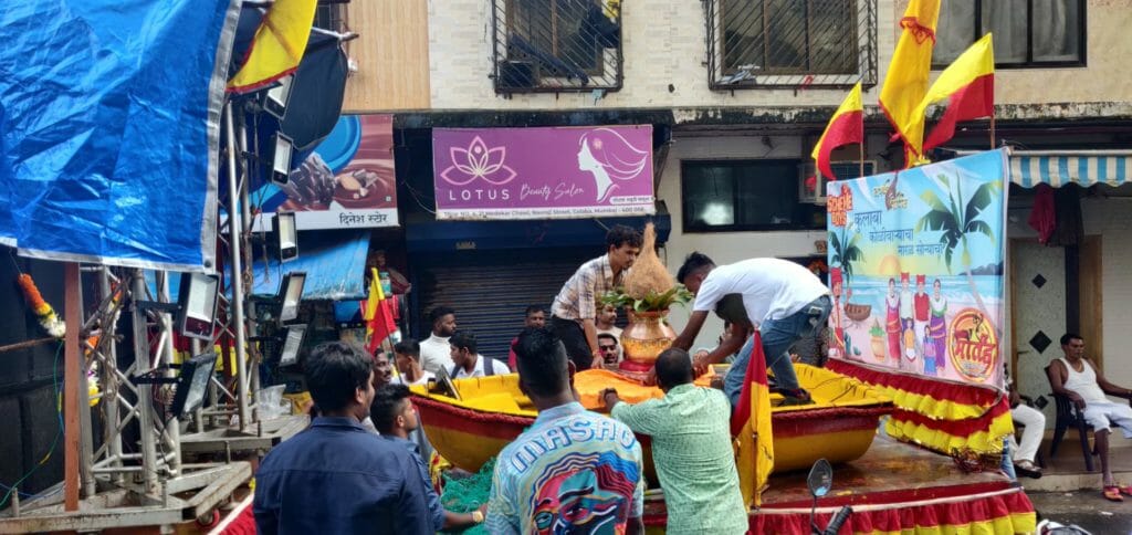 A group of young adults placing the coconut in the middle of the boat. There is banner in Marathi behind them that reads, "Colaba Koliwadyacha, Naral Sonyacha." (The golden coconut of Colaba Koliwada.)