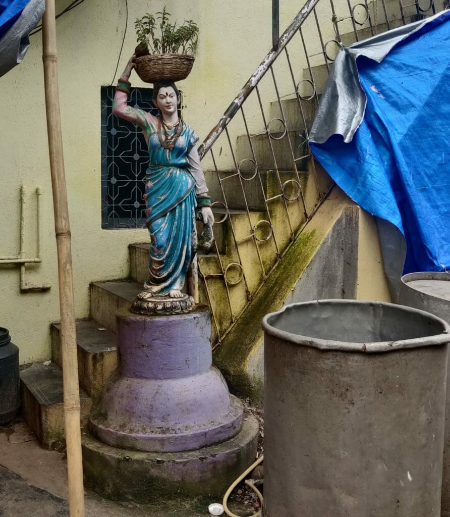 A sculpture of a Koli woman wearing a saree, her one hand is holding a fish and the other is balancing a basket. The sculpture is put up near stairs leading to a house.