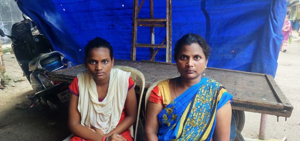 Priya and her mother Laxmi looking at the camera. They are sitting on two plastic chairs and there is a blue tarpaulin behind them.