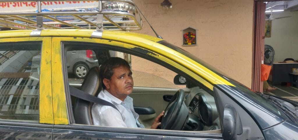 Sanjay Gupta in the driver's seat of a black and yellow taxi looking at the camera.