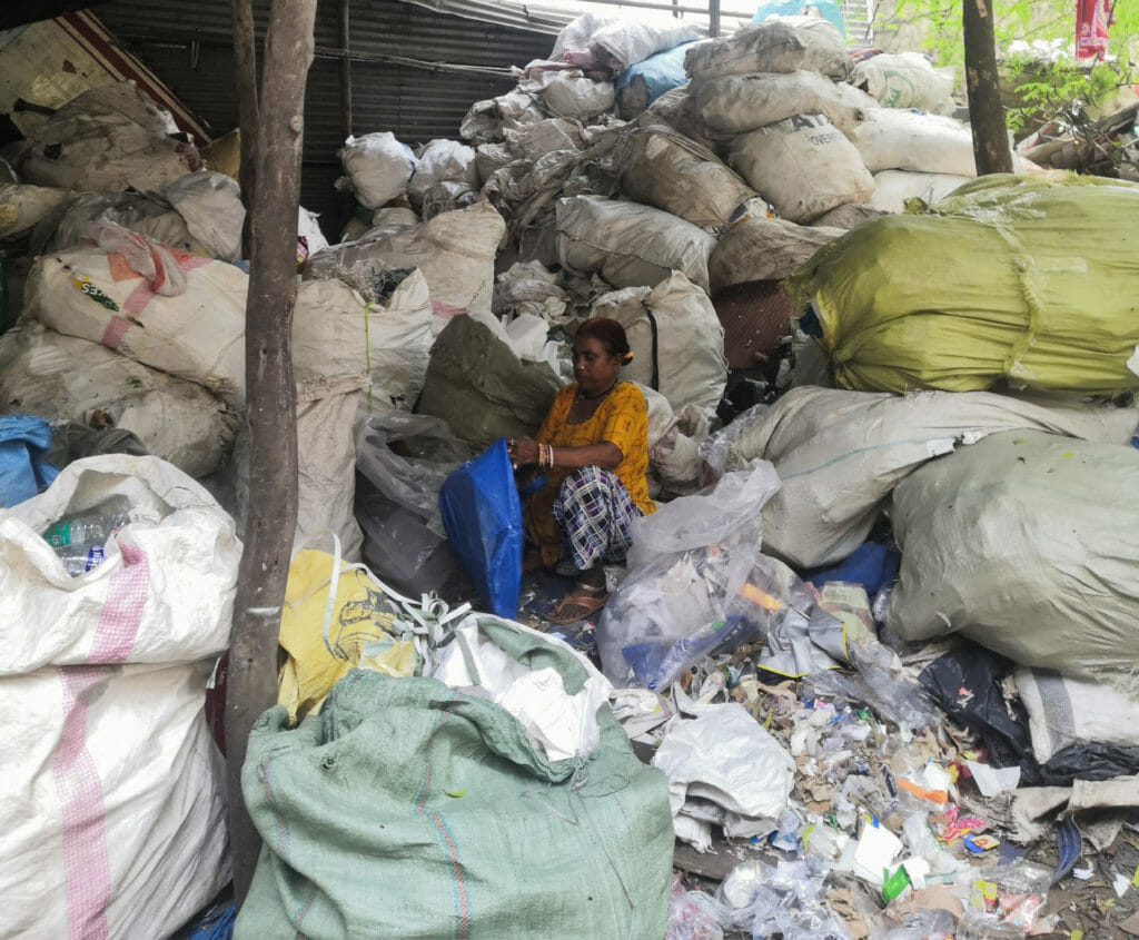 A woman sitting in the midst of large plastic sacks stacks up all around her, sorting waste from a bag
