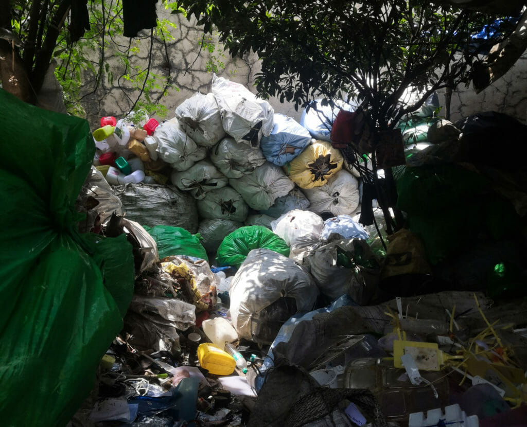 Plastic sacks filled with segregated dry waste stacked up on top of each other with other unpacked plastic waste and plastic cans around