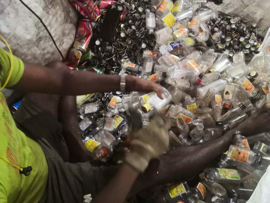 A man skinning the rings and caps off clear plastic bottles with a knife and gloves, separating the bottles and the rings and caps