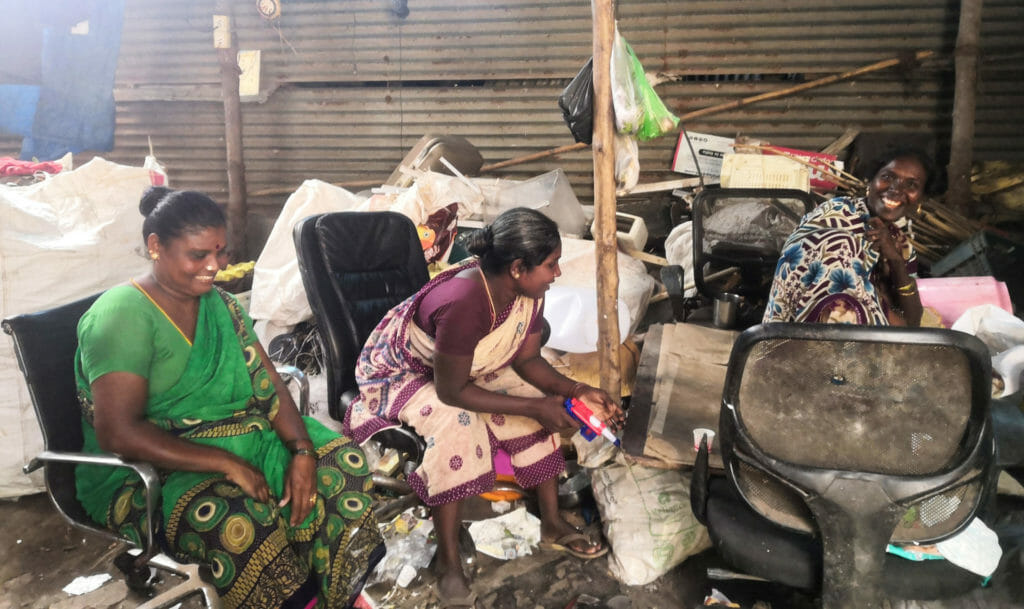 Three women sitting and laughing at the dry waste segregation centre on discarded office chairs and waste to be taken for recycling