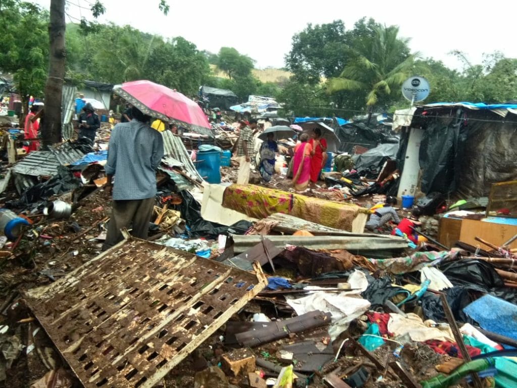 Houses, furniture and assorted items scattered around in a mess after the landslide in Malad in 2019, with people standing wtih umbrellas