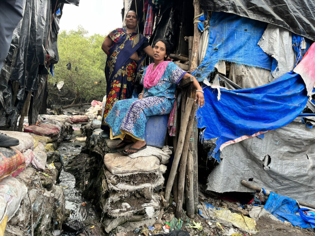 two women sitting on cement bags outside a home on stilts in a mumbai slum