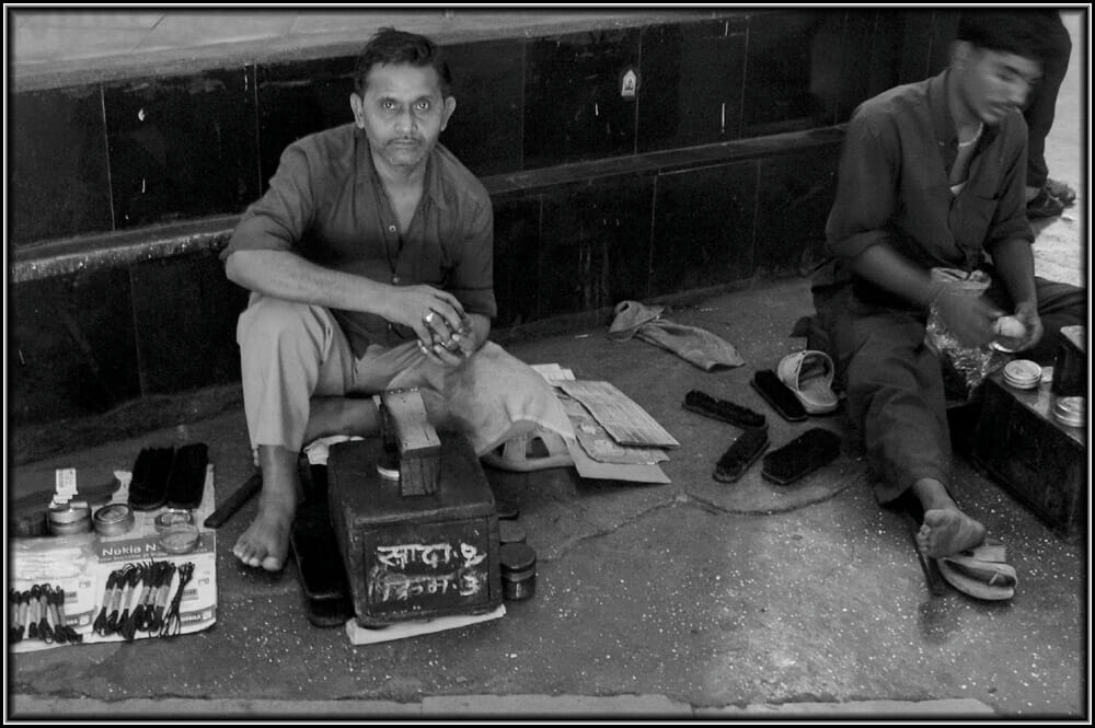 Two shoeshine workers sitting besides each other in front of their wooden platforms on a railway station