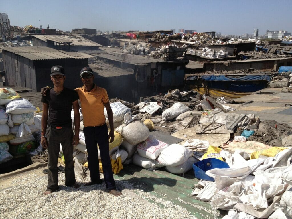 Two men standing on the roof of a slum, with bags around