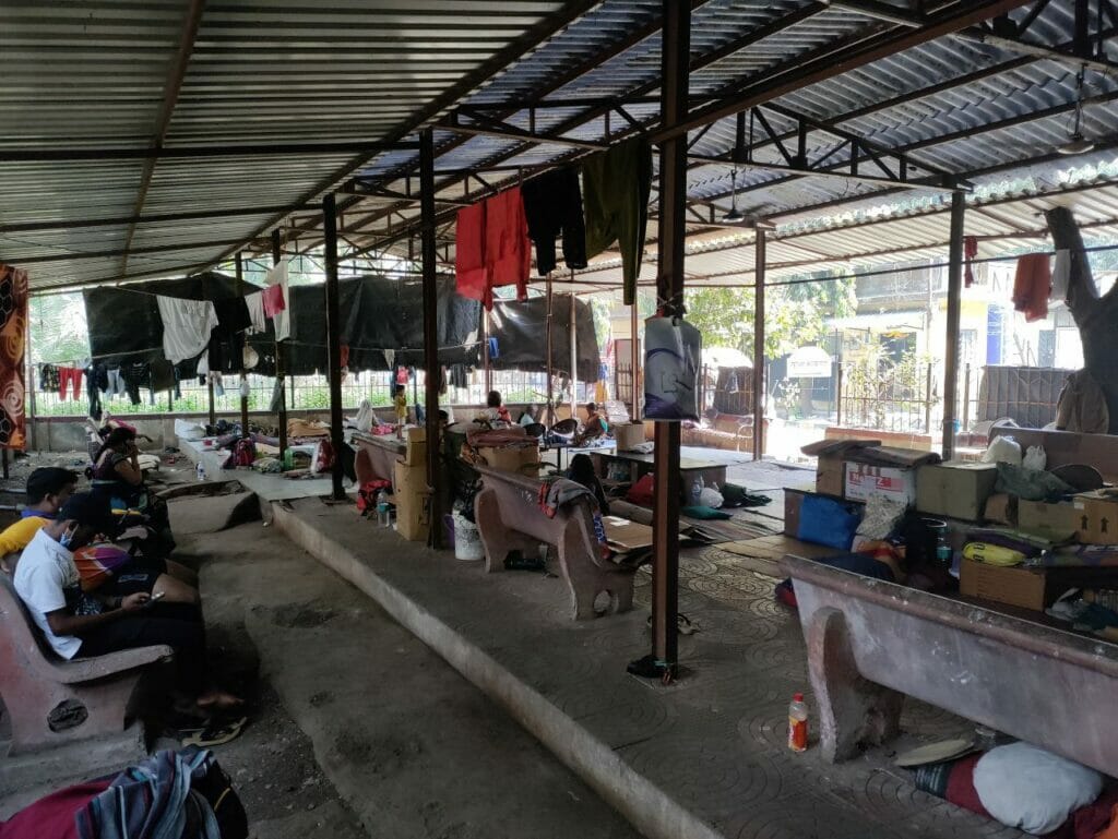Inside the shelter at tb hospital. There are clothes hanging on metal rods, benches for people to sit and other belongings of individuals.