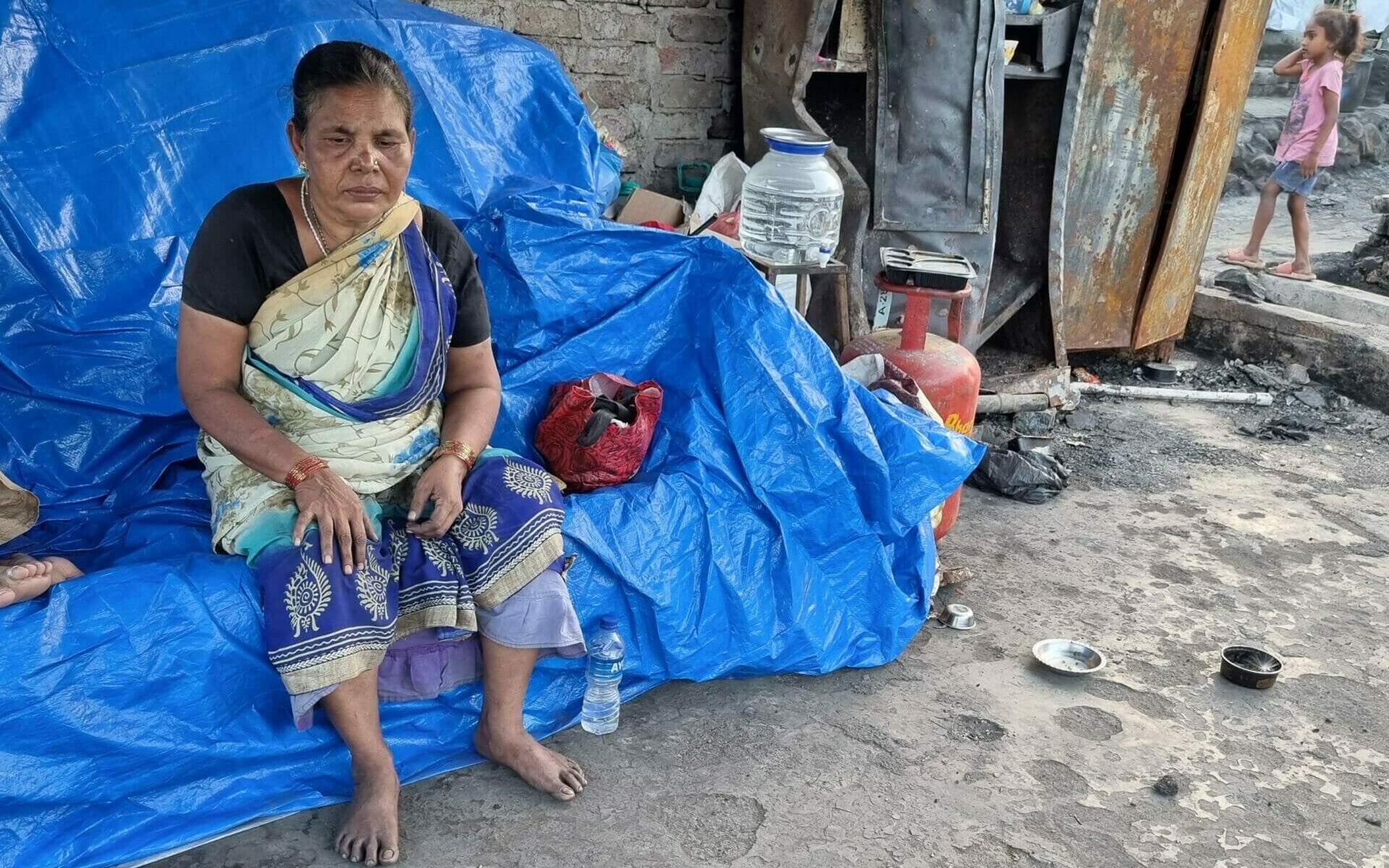 Sheela Ruke sitting on a tarpaulin sheet. There is a burnt metal cupboard next to her.