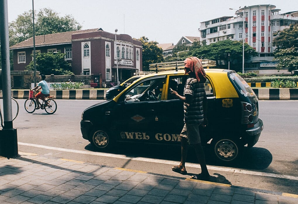 A man has covered his head with a cloth to avoid heat. He is walking next to Mumbai's black-and-yellow taxi.